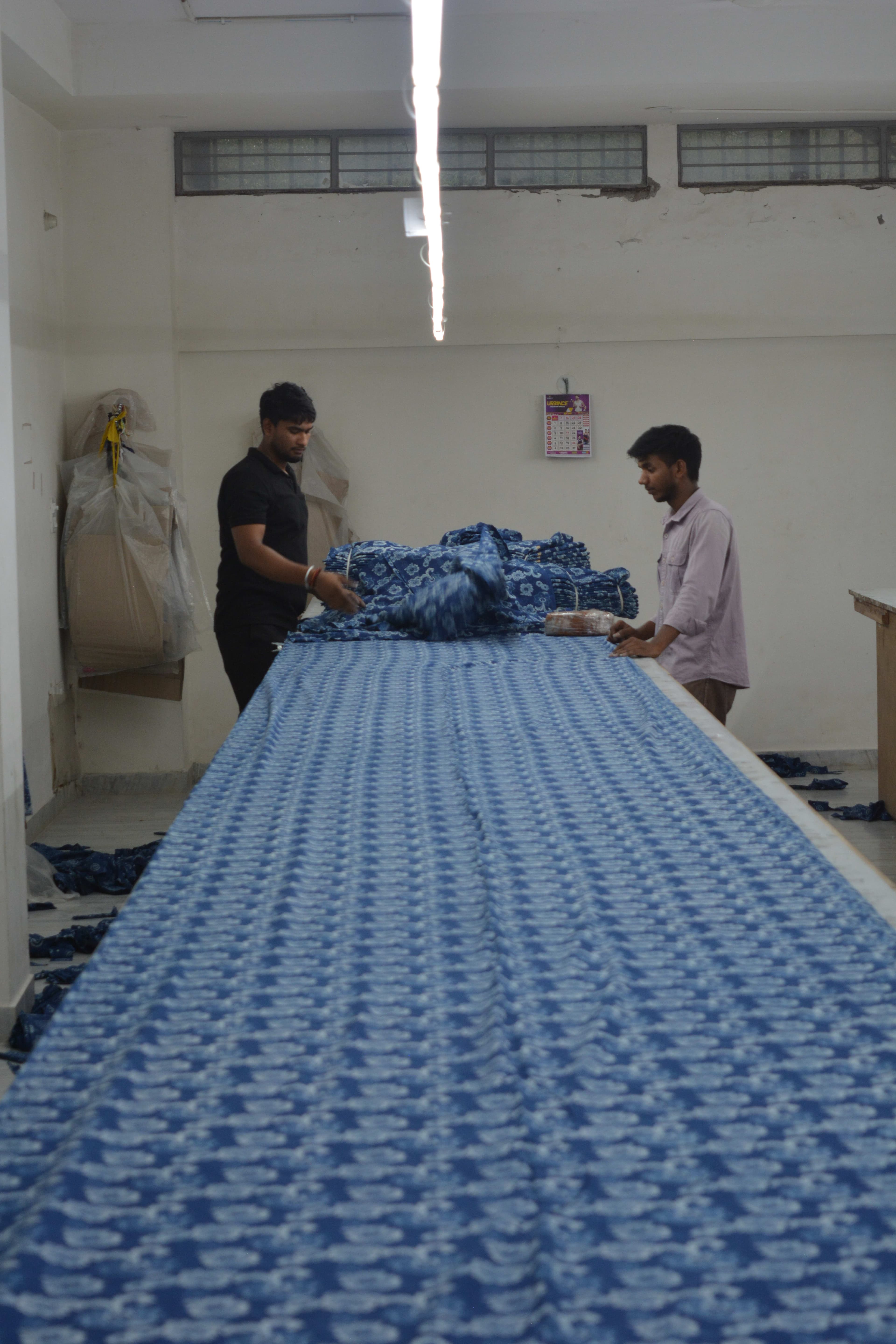 Two workers spreading blue printed fabric in layers across a long cutting table