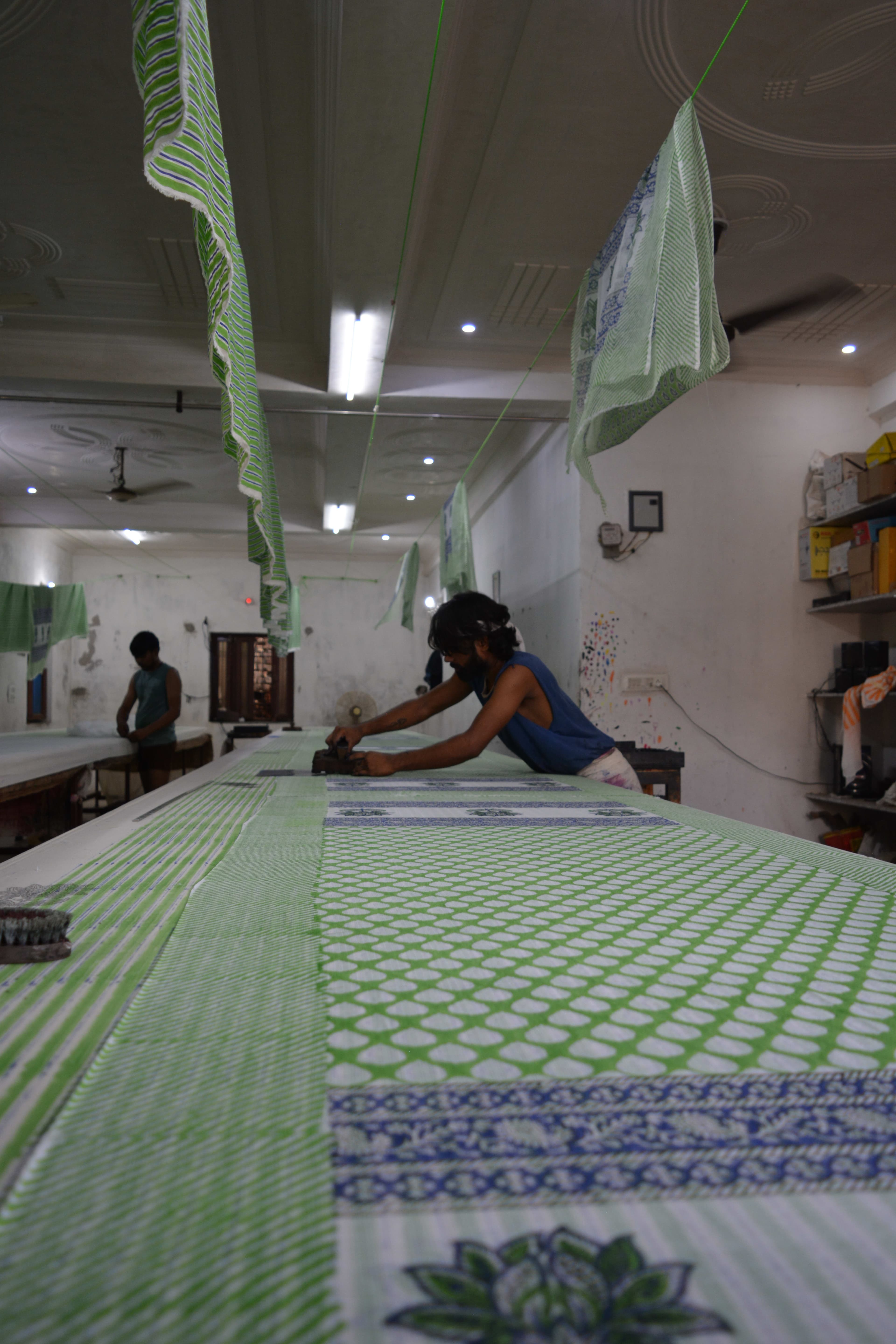 Low angle along the printing table showing the length of green and blue printed fabric, artisan working at far end