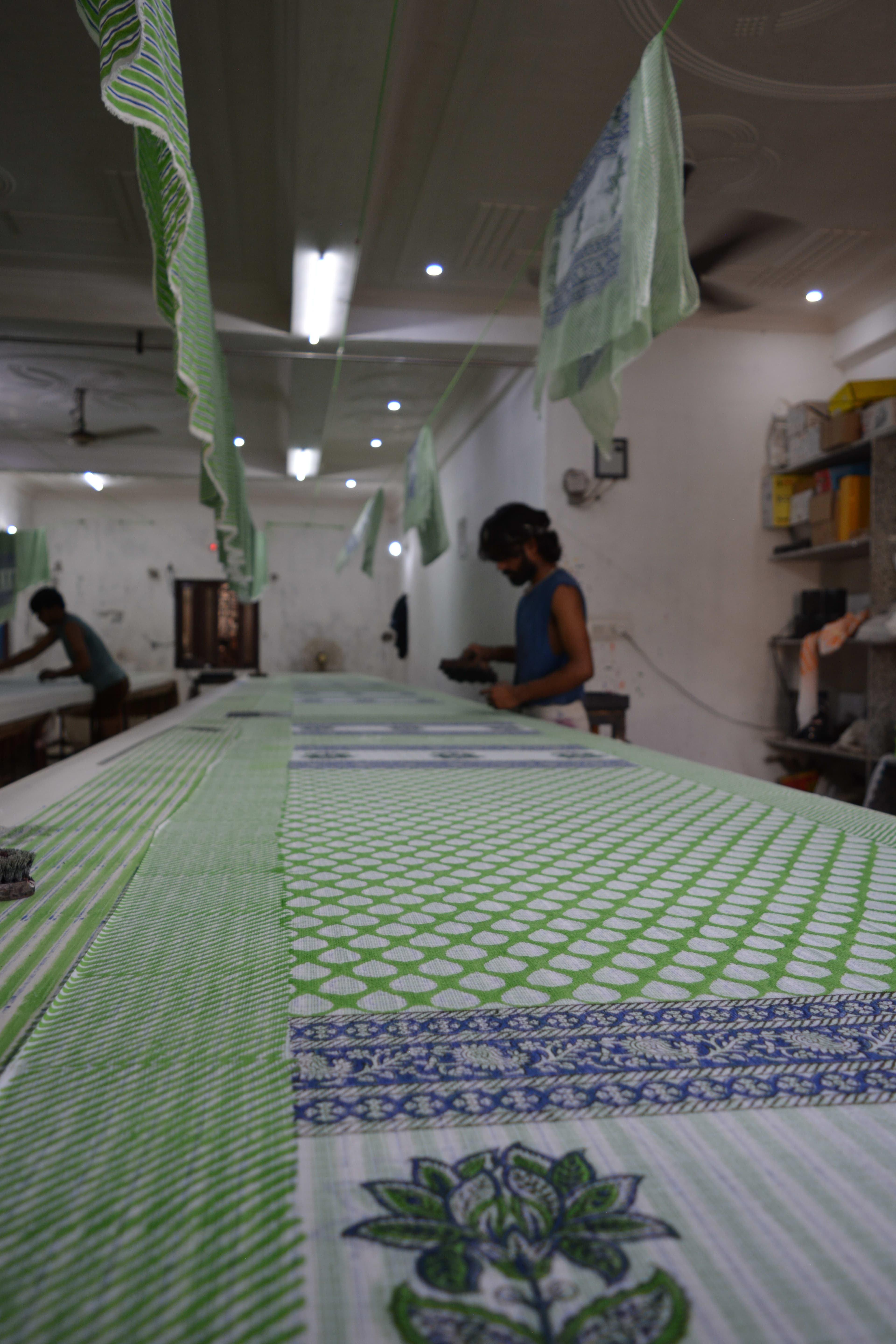 Artisan leaning over a long table adding a second colour pass to green printed fabric, printed pieces hanging to dry above