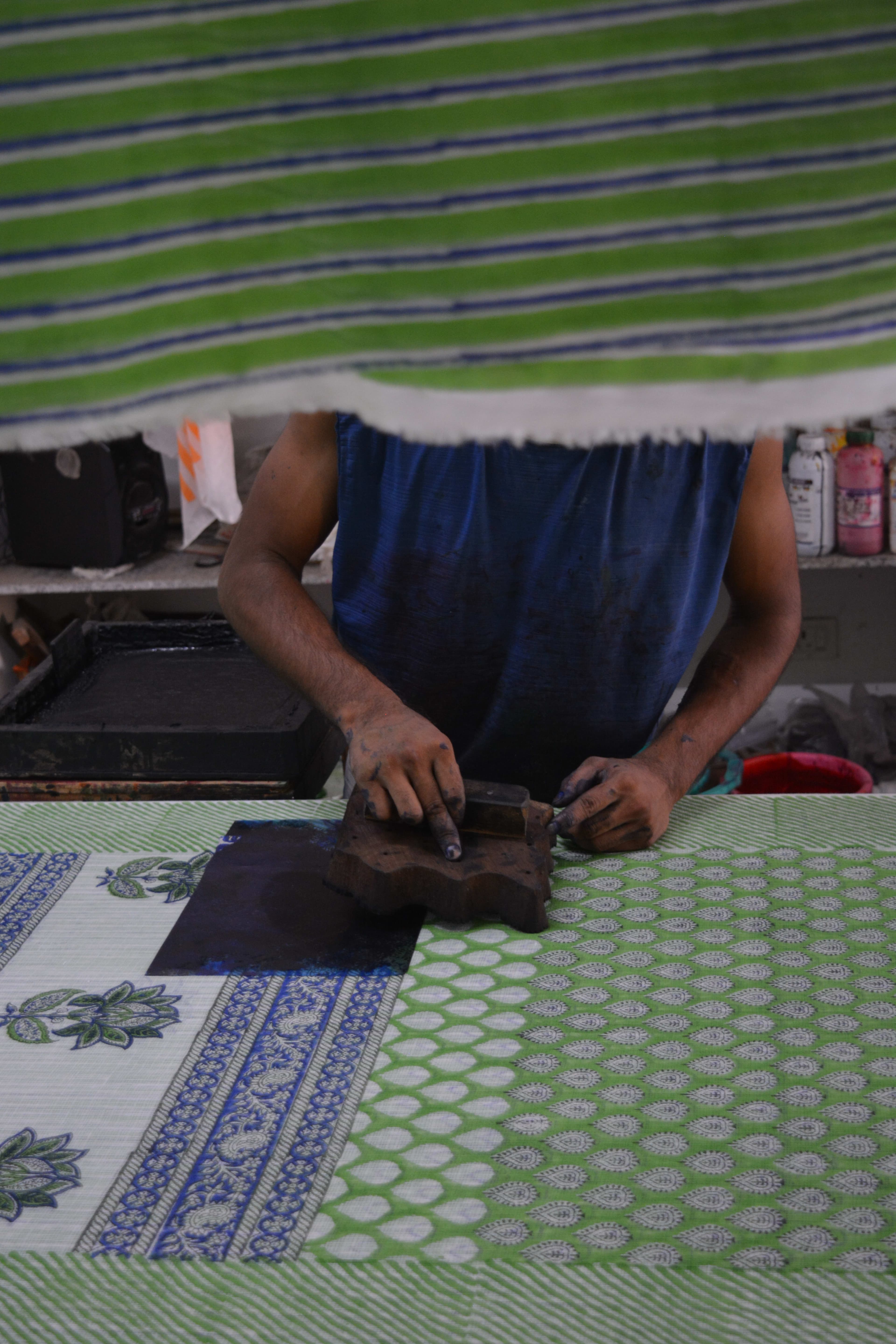 Artisan pressing a dark block onto multi-coloured printed fabric near dye tray, colour bottles visible behind