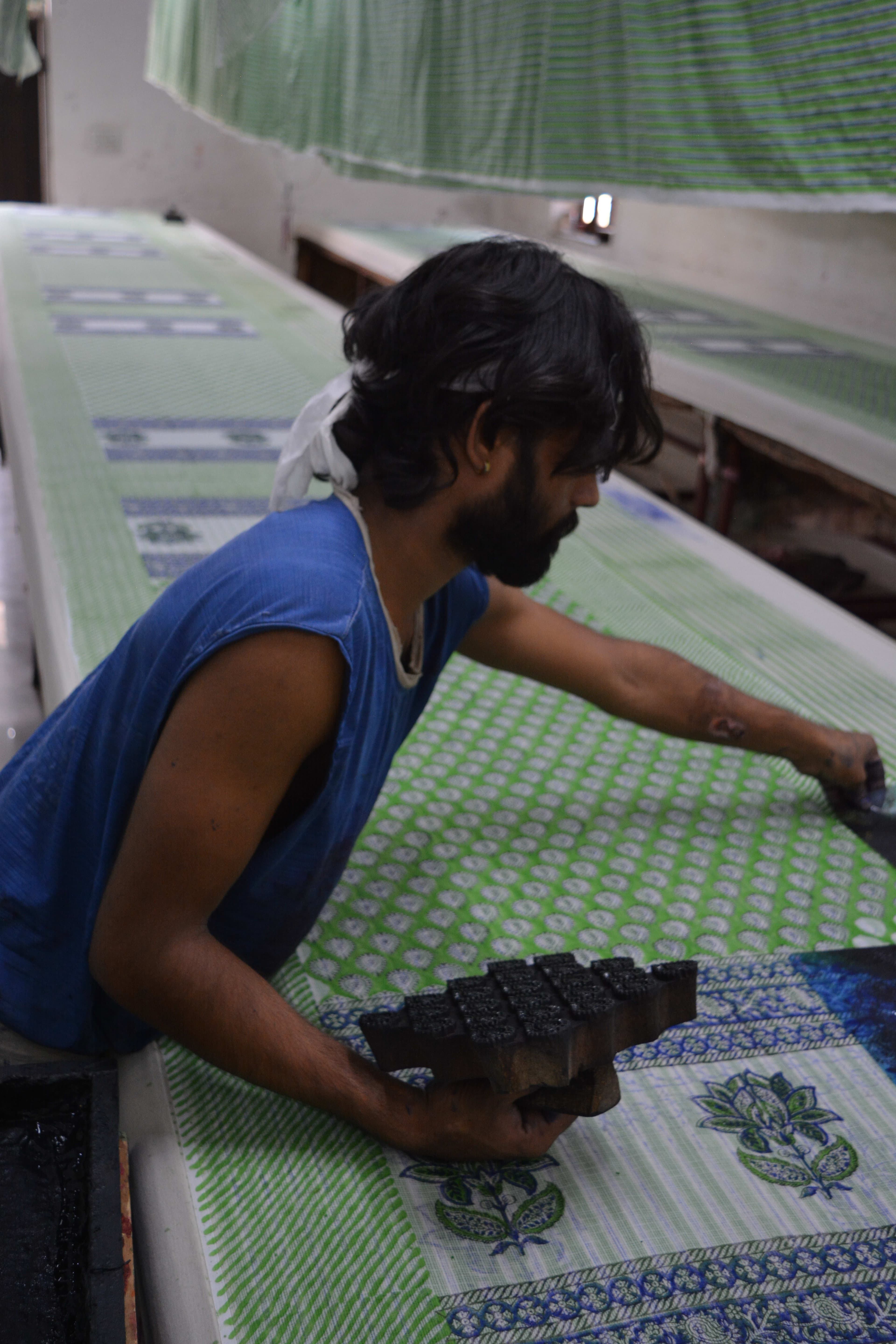 Artisan holding block mid-press against blue and green block-printed fabric, full printing room behind