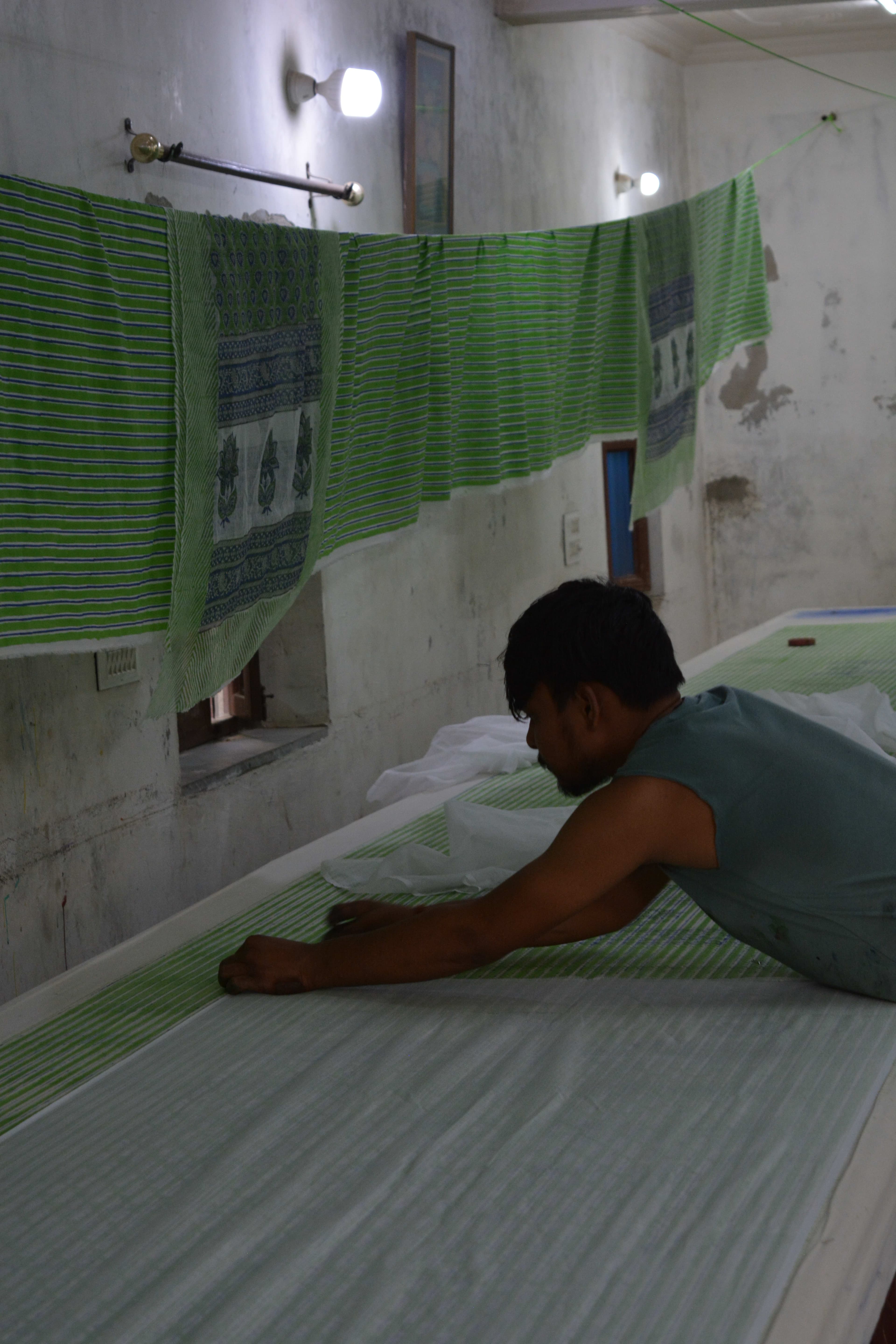 Artisan smoothing fabric flat on the printing table, printed pieces hanging to dry on the wall behind him